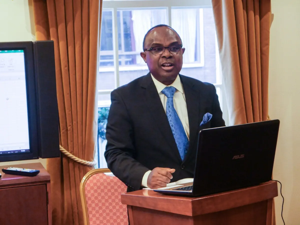 A speaker in a dark suit presents from a laptop at a wooden lectern.
