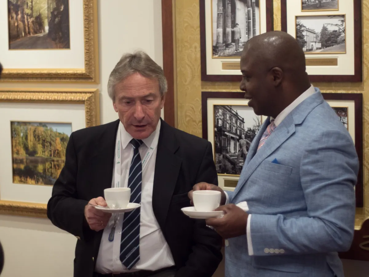 Two delegates in conversation over cups of tea in a room hung with framed photographs.