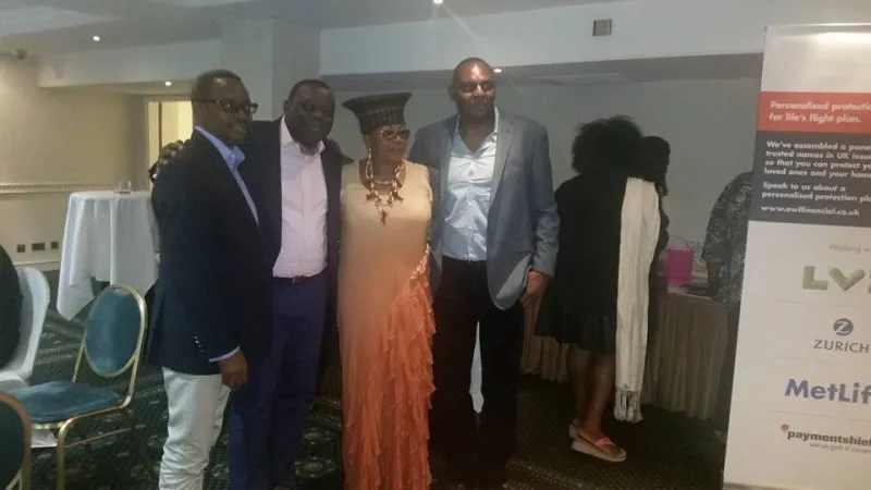 Four guests pose together, one in a gele headwrap and an orange gown, beside an exhibitor’s banner.