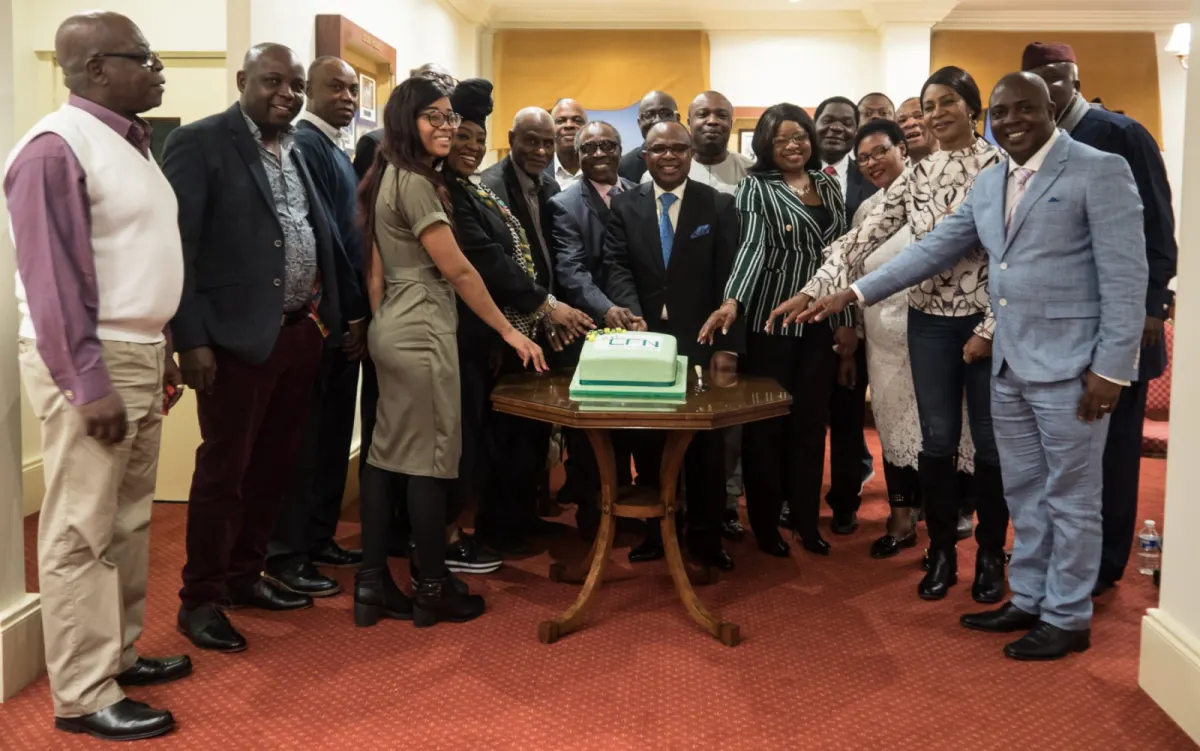 A large group in suits and Nigerian traditional dress gathers round a small table to cut a green-and-white celebration cake.