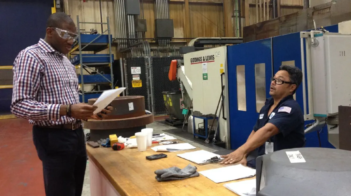 An engineer in safety glasses reads through a document with a machine operator at a workbench on a factory floor.
